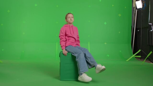 The girl sits on an imaginary parapet or roof. Teenager waving her legs while sitting in a virtual window on green screen background.