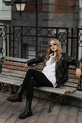 Fototapeta na wymiar Fashionable blonde woman model with black leather jacket and style sunglasses sitting on a bench at the city 