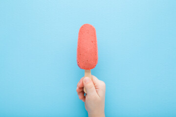Little child hand holding red strawberry ice cream with stick on light blue table background....