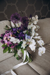 Wedding Floristics. Bridal bouquet of pink, white and purple flowers and greenery with white ribbon lies on the couch in the room