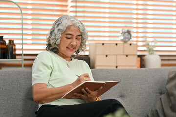 Relaxed middle aged woman sitting on couch and reading interesting book, enjoying leisure weekend time at home