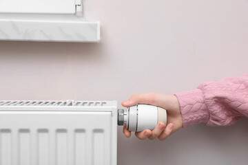 Girl adjusting heating radiator thermostat near white wall indoors, closeup