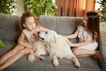 Beautiful purebreed dog, sand color American retriver lying on sofa with two kids, little girls in casual style clothes. Concept of happiness, family, care, animal and vet