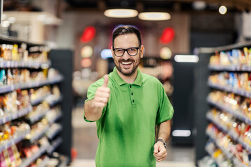 A happy employee is standing at the supermarket between the aisles and giving thumbs up at the camera. - obrazy, fototapety, plakaty