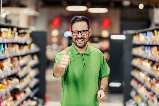 A happy employee is standing at the supermarket between the aisles and giving thumbs up at the camera.