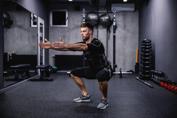 Fototapeta na wymiar Leg burning exercise, using new EMS technology. A young attractive man in EMS clothes in the gym doing squats with his arms outstretched. Electrical muscle stimulation, strong movement
