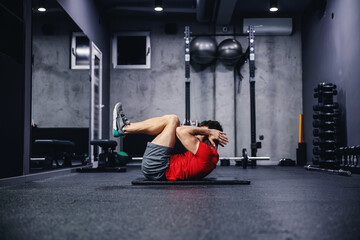 Fototapeta na wymiar A young handsome sportsman exercises, doing abdominal crunches. He does sit-ups in the fetal position, wearing sportswear indoor gray sports studio. Weight loss and healthy living