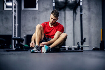 Fototapeta na wymiar Preparation for strong functional training for the whole body. Pause between exercises. A handsome and hot sporty man ties a shoelace on his sneakers in the gym with exercise equipment