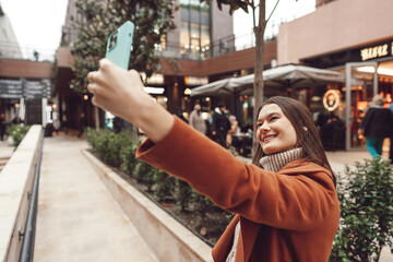 Naklejka premium A happy woman in a coat standing on the street and taking a selfie