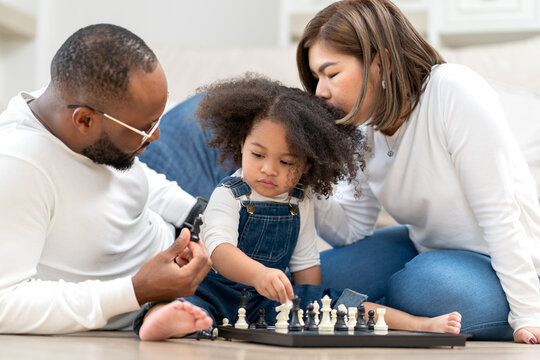 Cute little girl playing chess board as her father sitting by. Multiracial family happy leisure together with daughter kid at home. Multiethnic child has fun playing chess pieces with her dad and mom. - Powered by Adobe