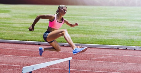 Caucasian female athlete jumping over a hurdle against sports field in background