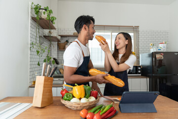 Young Asian couple in aprons learning to select ingredients Bread that is suitable eating in morning along with salads, vegetables, fruits via online tablets lifestyle and healthy cooking concept