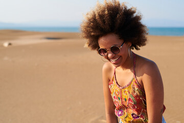 Beautiful modern ethnic woman with Afro hairstyle wearing colorful top and sunglasses smiling on beach