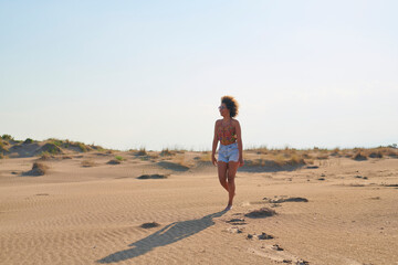 Full length portrait of happy beautiful woman with afro hairstyle in sunglasses, top and denim shorts walking in sunny desert on summer day. She is looking away with excitement.