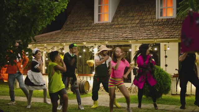 Group of funny , stylish multi aged dancers dancing near village house at night . Kids dancing with house workers . Multi ethnic people having fun . Beautiful background with different lights .