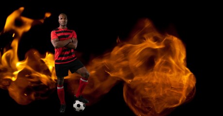 Composition of male football player standing with ball over flames on black background