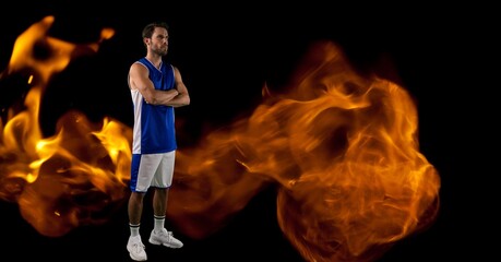 Composition of male basketball player standing with arms crossed over flames on black background