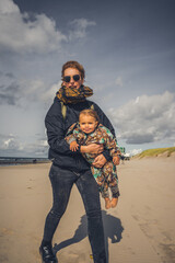 Naklejka premium Mutter mit Kleinkind auf dem Arm ab einem Strand an der Ostsee, Wetter ist frisch und der Wind geht stark, trotzdem sind beide fröhlich und lachen 