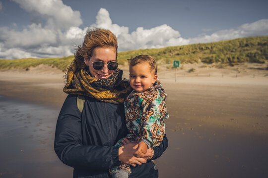 Mutter mit Kleinkind auf dem Arm ab einem Strand an der Ostsee, Wetter ist frisch und der Wind geht stark, trotzdem sind beide fr&ouml;hlich und lachen 