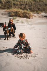 Kleinkind spielt im Sand am Starn, im Hintergrund hockt Mutter mit Boxer Hund am Rand der Dünen