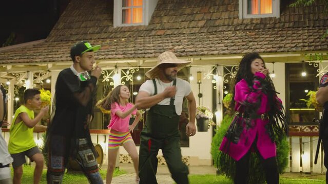 Group of funny , stylish  multi aged dancers dancing near village house at night . Kids dancing with house workers . Multi ethnic people having fun . Beautiful background with different lights .
