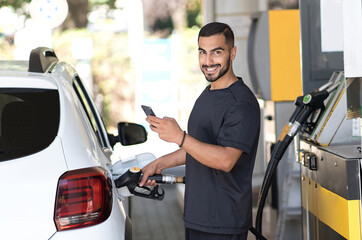 Positive man refueling his luxury car and messaging smartphone at the gas station