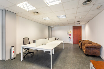 Large conference room with tiled floor and bright lamps on the unfashionable ceiling. Along the walls are outdated armchairs with worn upholstery.