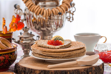 Russian traditions. Russian holiday Maslenitsa. Still life with a cup of tea, a stack of pancakes, red caviar, lemon, bagels, a lollipop cockerel, wooden spoons (khokhloma) and samovars.