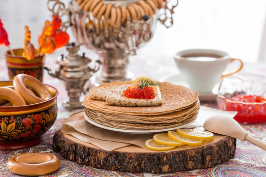 Russian traditions. Russian holiday Maslenitsa. Still life with a cup of tea, a stack of pancakes, red caviar, lemon, bagels, a lollipop cockerel, wooden spoons (khokhloma) and samovars.