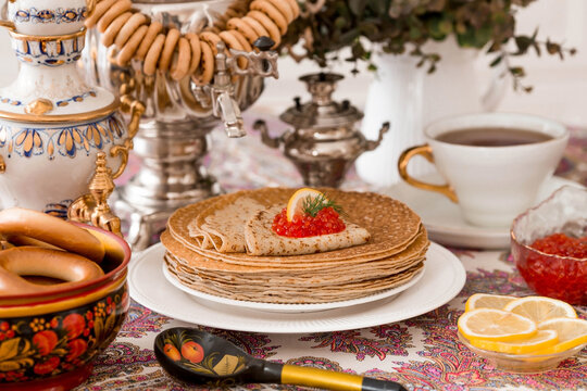Russian traditions. Russian holiday Maslenitsa. Still life with a cup of tea, a stack of pancakes, red caviar, lemon, bagels, a lollipop cockerel, wooden spoons (khokhloma) and samovars.