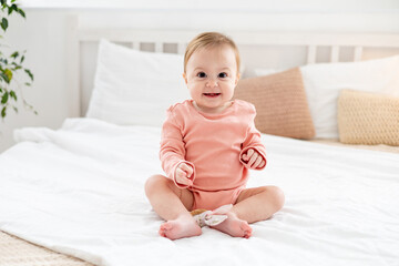 a baby girl on a bed in a bright bedroom in a pink bodysuit smiling or laughing