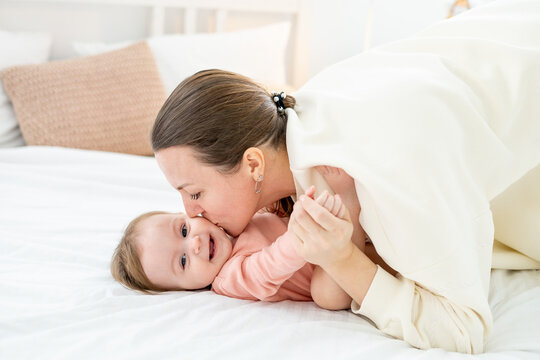 mom gently kisses and hugs her little daughter at home on the bed and have fun together, family motherhood and love for the baby