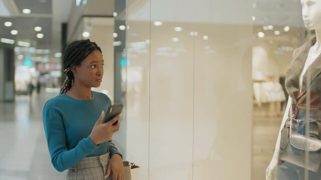 African American young woman taking photo of clothes on mannequin on her smartphone during shopping in the mall