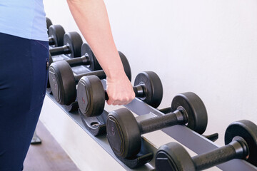 Naklejka premium Woman in the gym takes a dumbbell from the rack. Black metal dumbbells of different weights on a rack in a gym. Sports equipment for increasing muscle mass. The concept of doing sports.