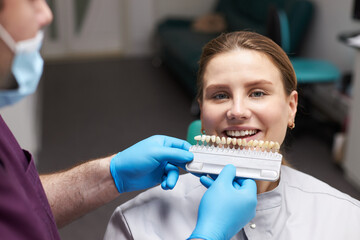 Dentist placing color chart, over a woman's teeth, choosing veneers shade, according to Vita scale