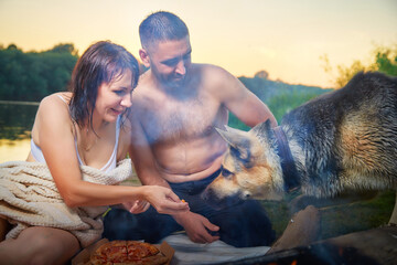 Happy wet couple relaxin, having fun and hugs with big dog near fire in camping on nature at summer...