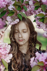 Fototapeta premium Pretty young girl with closed eyes and hair strands on her face in a frame of blooming sakura tree branches with gentle pink flowers dreaming in spring park on sunny day of springtime, vertical