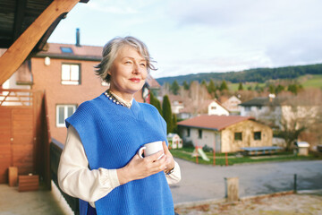 Portrait of beautiful 50 -55 year old woman posing on balcony, holding cup of tea or coffee