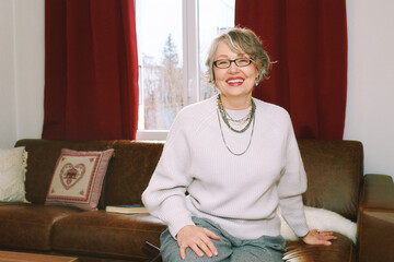 Interior portrait of 50 - 55 year old woman sitting on couch , wearing glasses