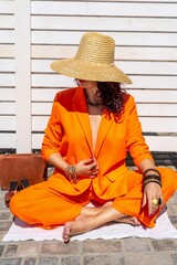 Stylish woman in an orange suit with a hat sits on a rug on a white striped background. On the hands are jewelry rings and bracelets, sandals and a bag stand side by side.