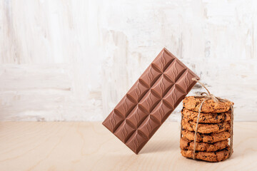 A stack of chocolate chip cookies tied with twine and a bar of chocolate on a on a wooden table.