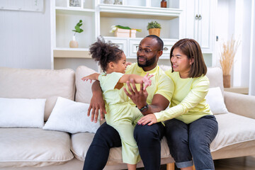 Young family with small daughter sitting on sofa playing and laughing together in the living room