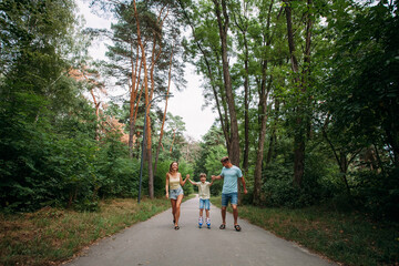 Fototapeta premium A happy young family, dad, mom and son are skating hand in hand with their parents, on roller skates, along a path in the park. The whole family is dressed in shorts and T-shirts.