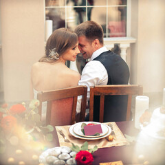 Beautiful smiling newlyweds in stylish outfits are sitting at the wedding table set against the...