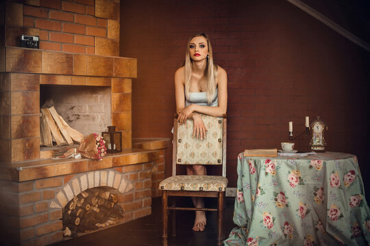 A gorgeous girl with bright make-up, with blond hair, in a dress, stands in a room with a fireplace, near the table. There is a candlestick with candles, a clock, a cup and a book on the table.