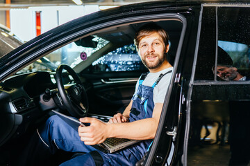 Cute smiling mechanic or electrician sitting in car and using laptop at car service. Vehicle wiring inspection. Car service, repair, maintenance and people concept.
