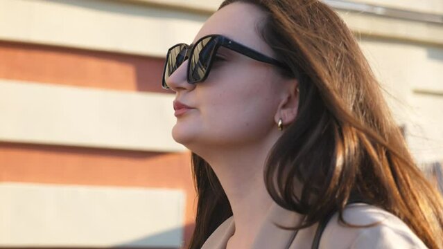 Profile of young successful businesswoman in sunglasses walking at sidewalk. Attractive brunette girl going along urban street. Happy beautiful lady commuting to work. Close up Slow motion