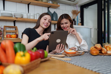 Beautiful woman and friend talking and using laptop at kitchen room. Girl friends couple living together at home.