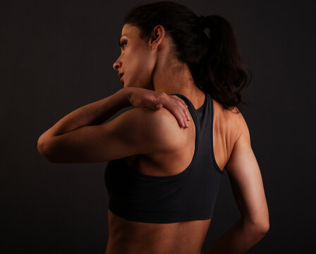 Female sporty muscular with ponytail doing stretching workout of the shoulders, blades in sport bra, holing the neck the hand on dark grey background with empty copy space. Back view.