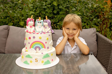 Cute five years old child, sitting in garden with big homemade cake with unicorns, celebrating birthday